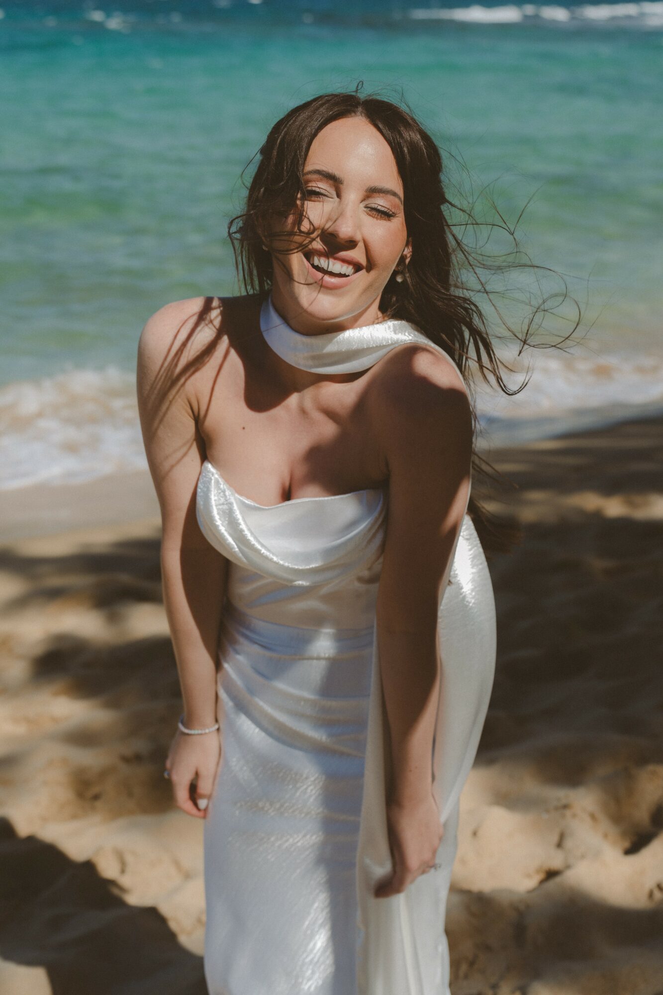 Smiling woman in white dress on beach with ocean background, hair blowing, sunlight, sandy shore, relaxed pose