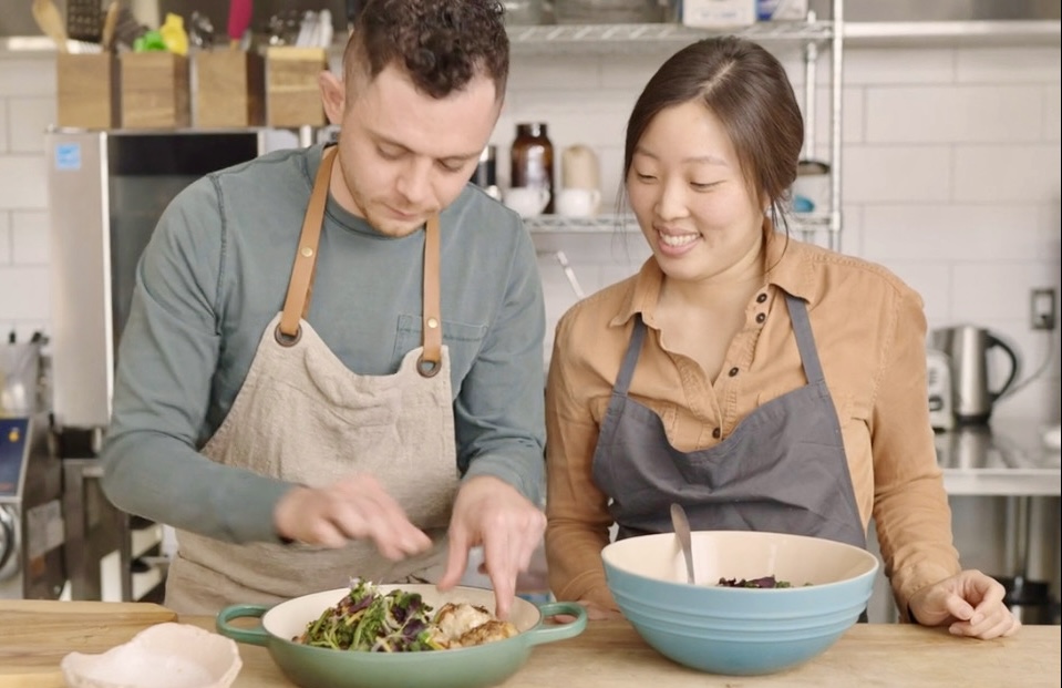 Two people preparing food in a kitchen, smiling, with bowls and ingredients on the counter.