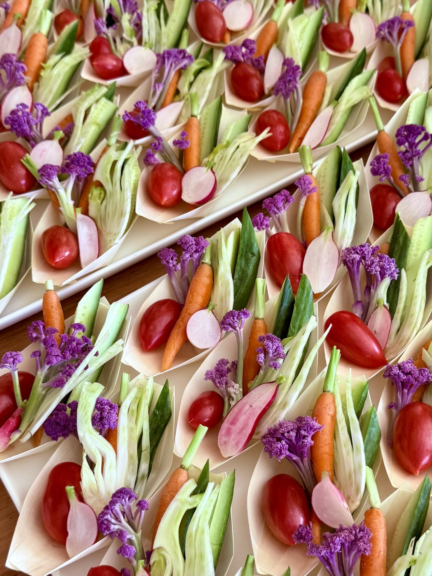 Colorful appetizer bites with cherry tomatoes, green vegetables, purple flowers, and small breadsticks on white trays.