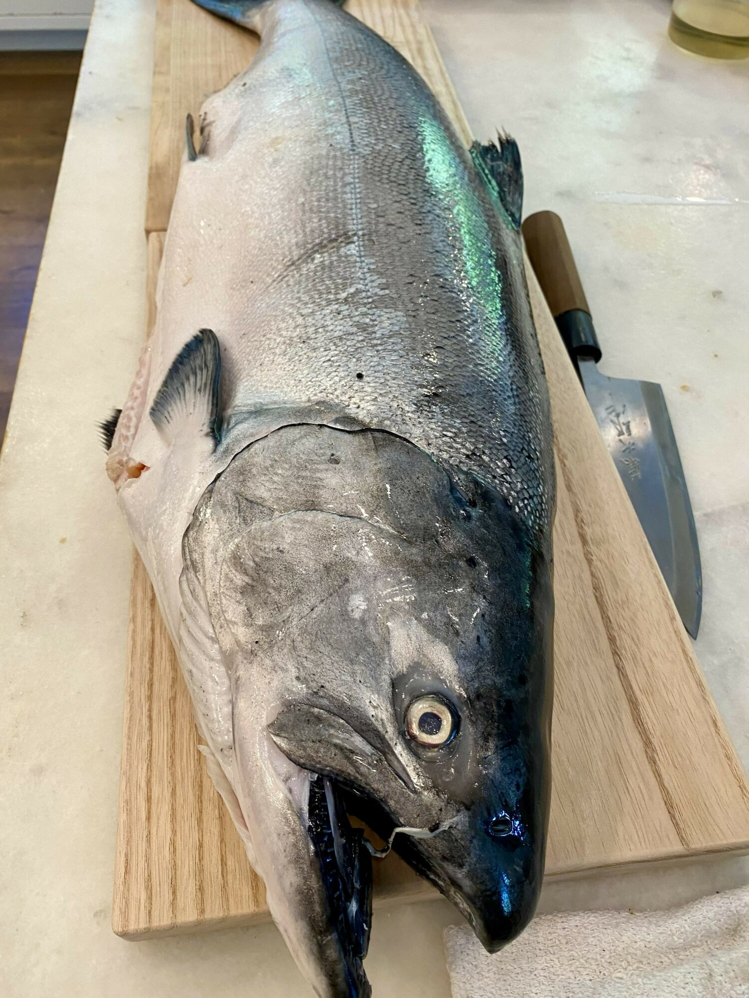 Fresh fish on a wooden cutting board with a knife nearby, showing silver and dark coloration, open mouth, and visible eye.