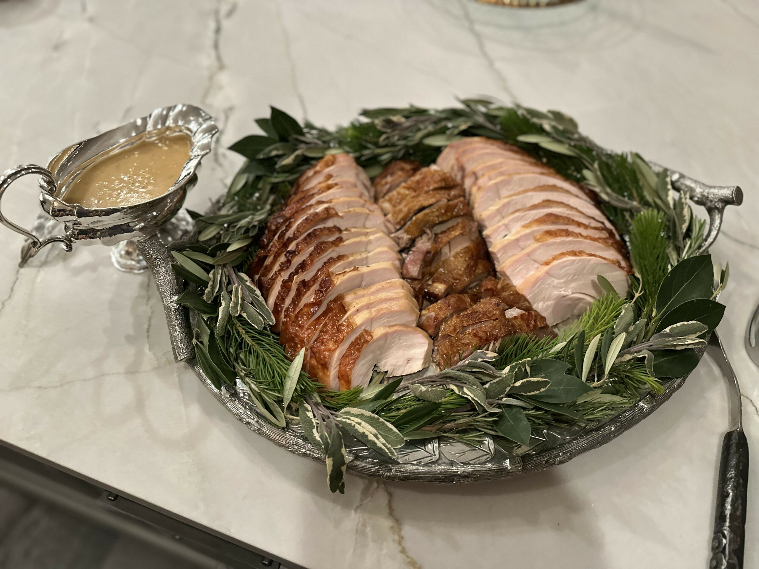 Sliced cooked meat on a decorative silver platter with green leaves, accompanied by a small silver sauce container, on a white surface.