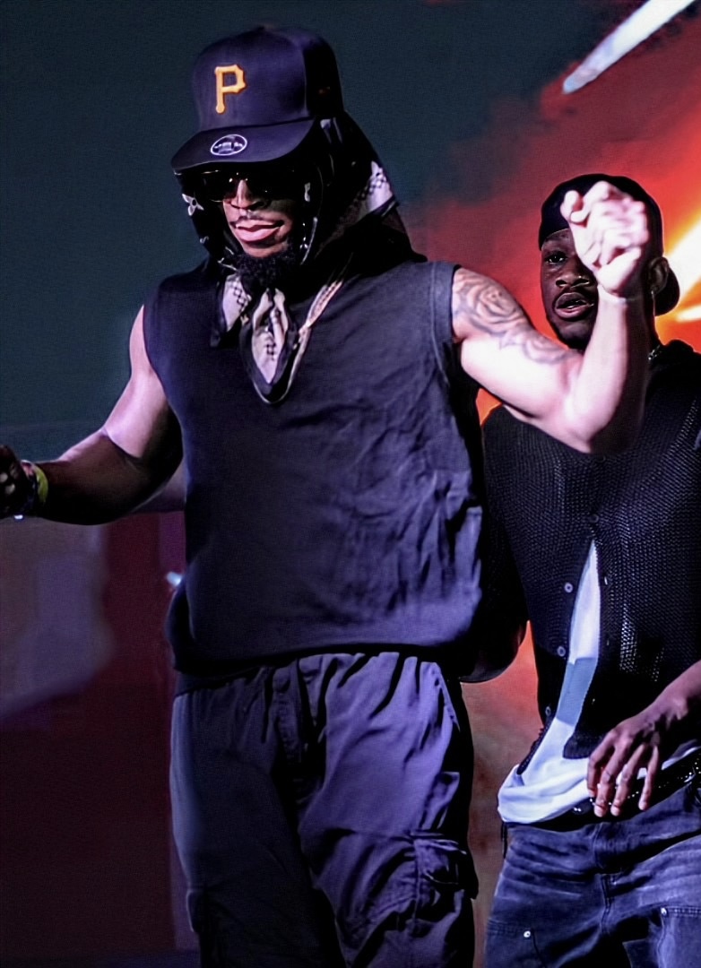 Person wearing a black cap with a yellow 'P', sunglasses, sleeveless shirt, and dark pants, raising a fist.