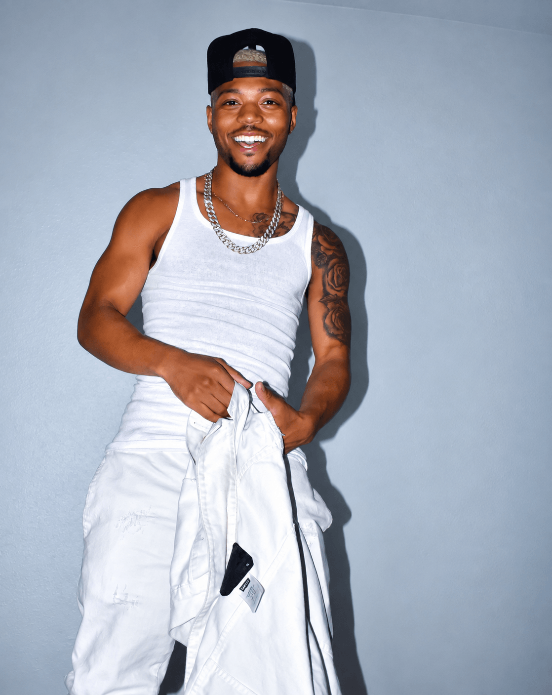 Smiling man in a white tank top, white pants, black cap, and jewelry, standing against a light blue background.