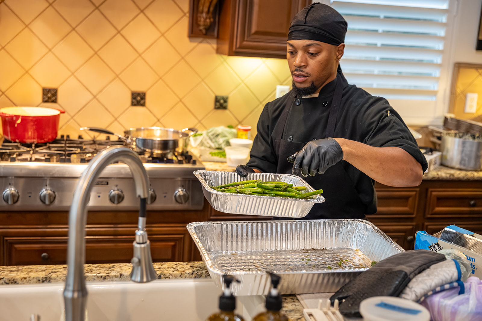 Chef in black uniform and head covering preparing food in a kitchen with wooden cabinets and tiled backsplash.