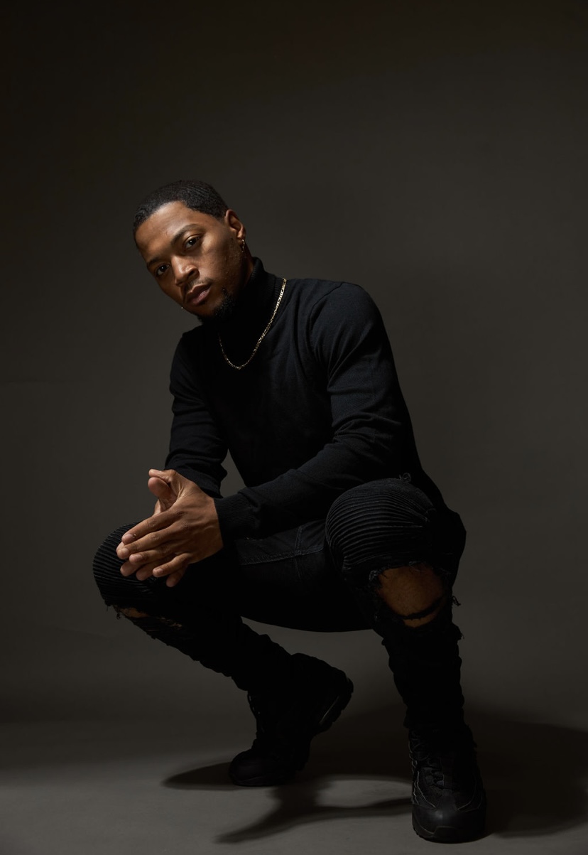 Person crouching with hands clasped, wearing black clothing and boots, against a dark background.