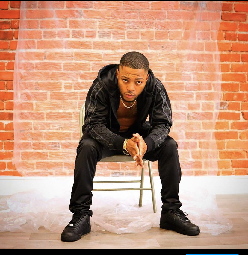 Young man sitting on a chair against a brick wall, leaning forward with hands clasped, wearing black clothing and sneakers.