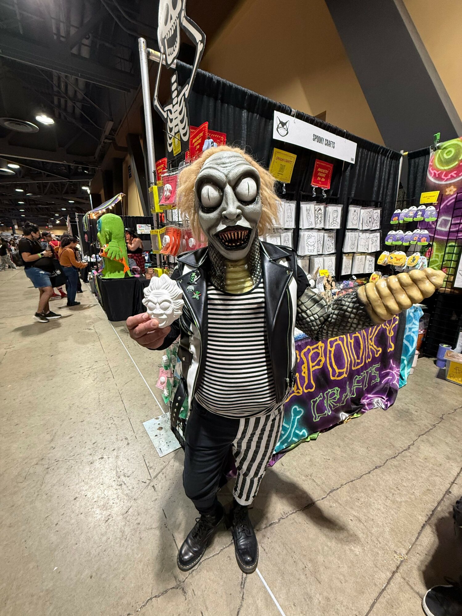 Person dressed as a skeleton with a skull mask, striped shirt, and black pants, standing in a store aisle with colorful products.
