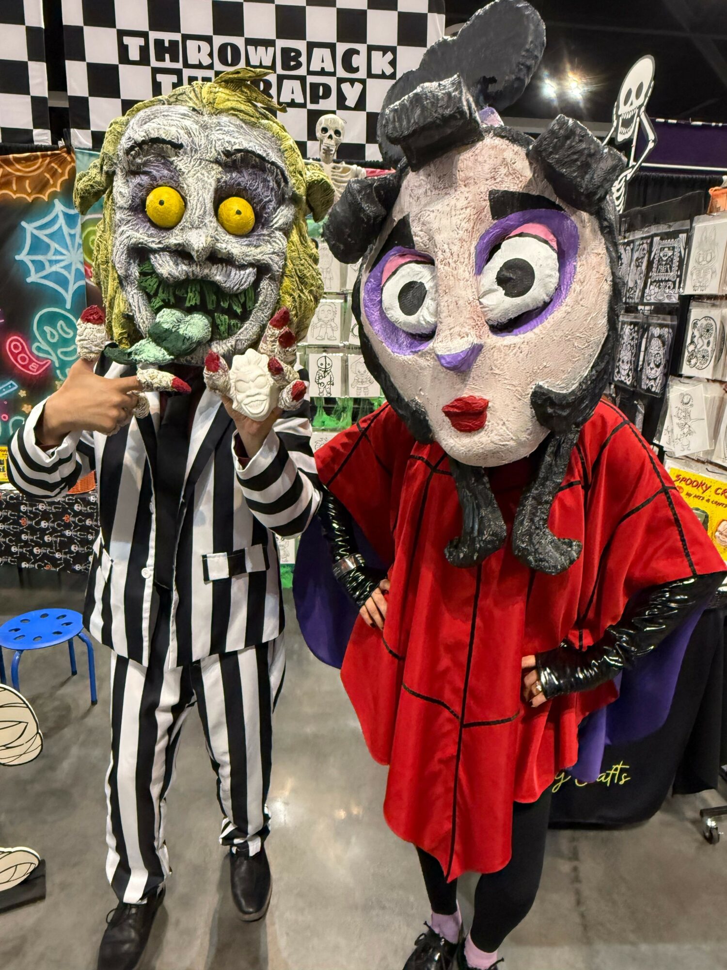 Person in a striped costume with yellow eyes and a skull face, holding a phone, standing next to a person in a vampire costume with purple eyes and red cape, in a store.