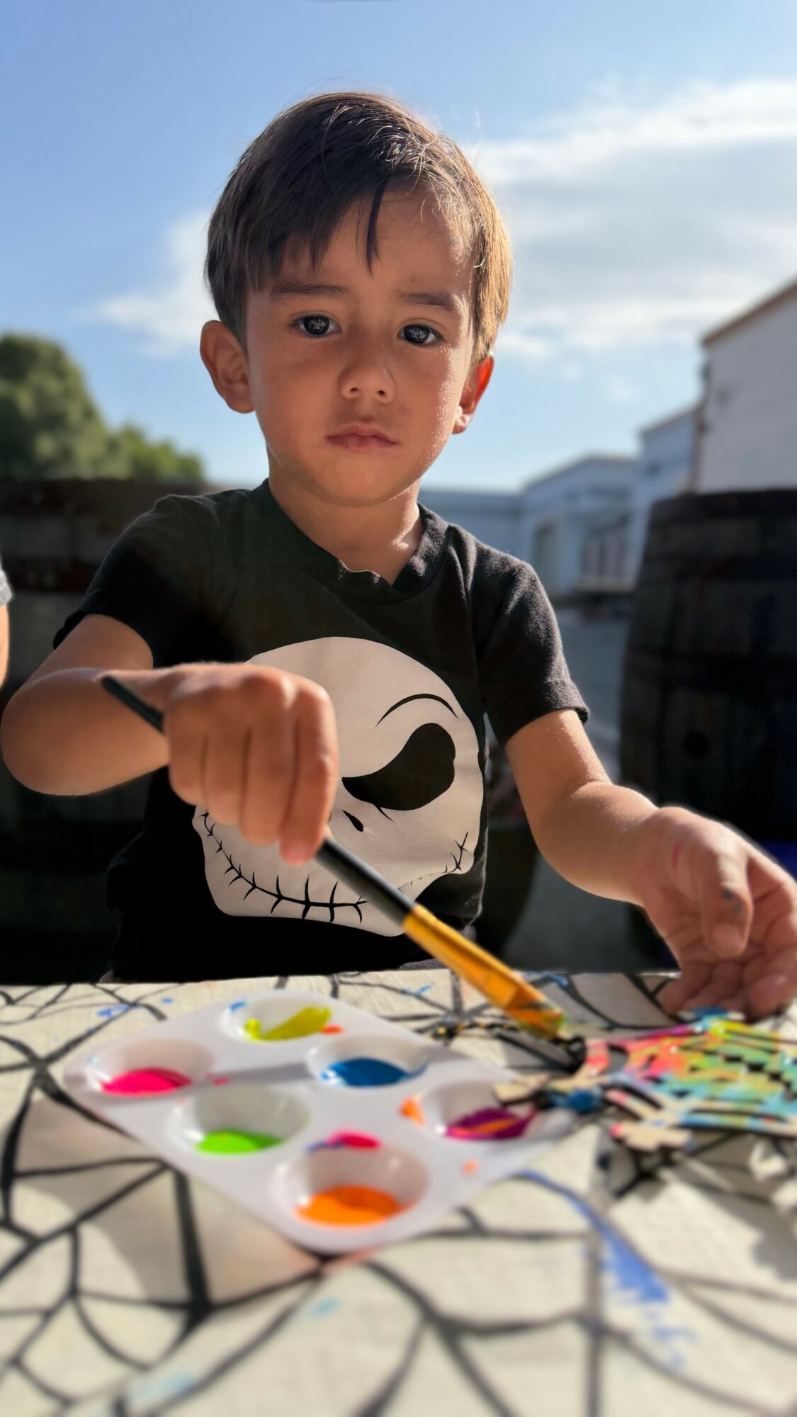 Young boy with dark hair and a black shirt with a skull design, painting outdoors on a sunny day.