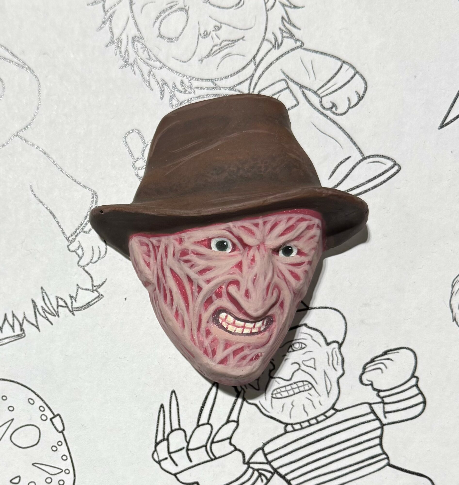 A 3D mask of a person's face with an angry expression, wearing a brown hat, placed on a drawing background.