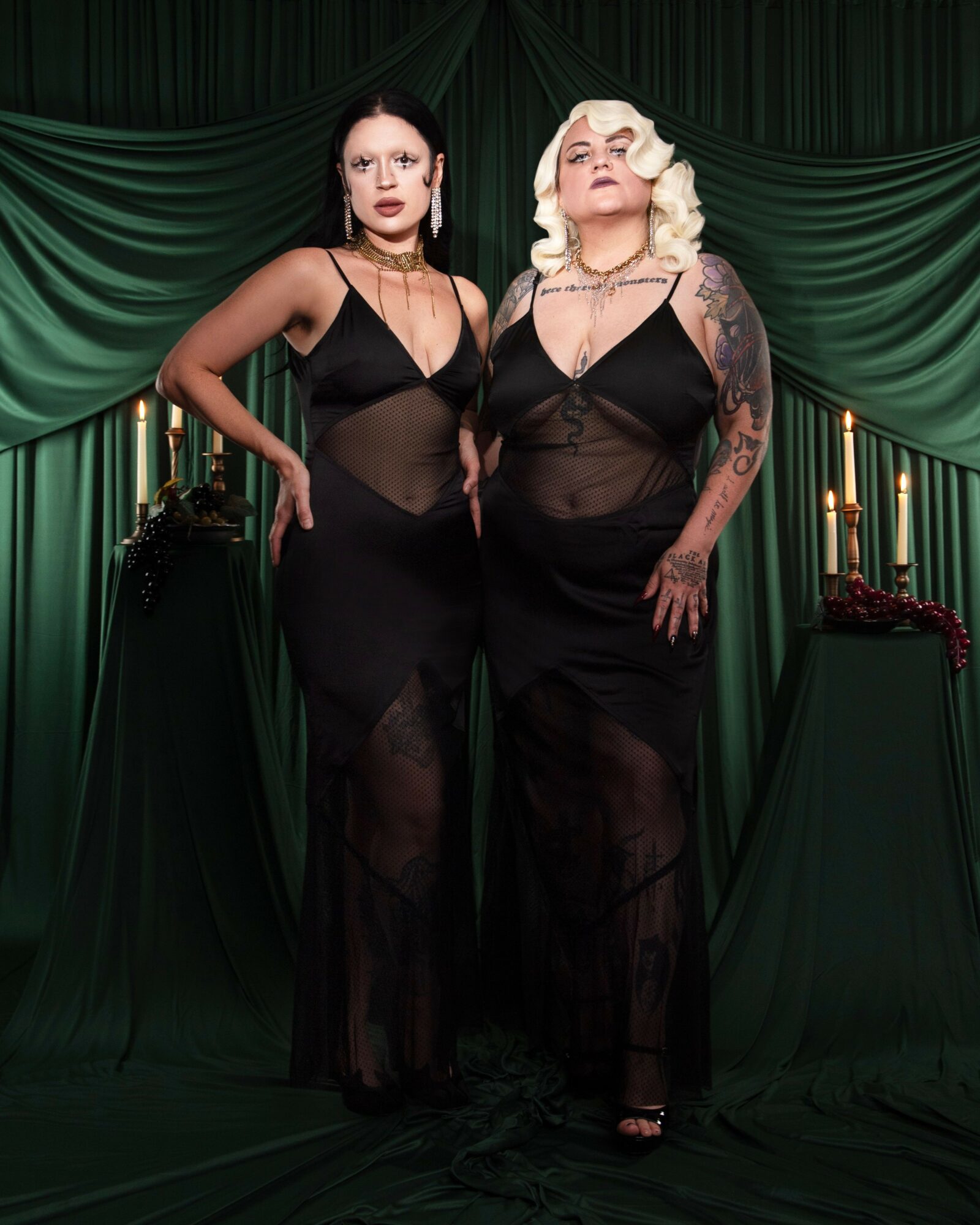 Two women in black dresses stand in front of green curtains with candles on tables, posing confidently.