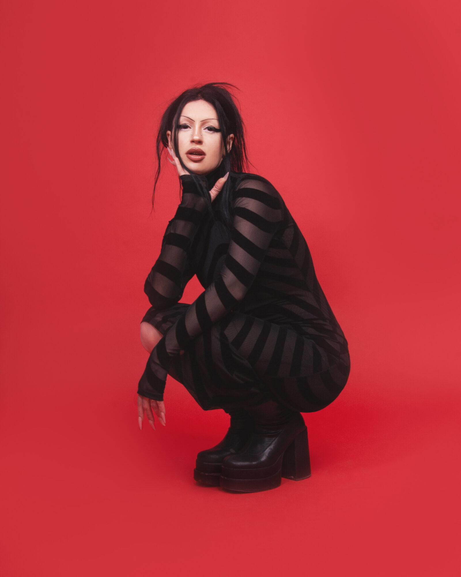 Woman crouching in black striped outfit against red background, with dark hair and makeup, looking at camera.