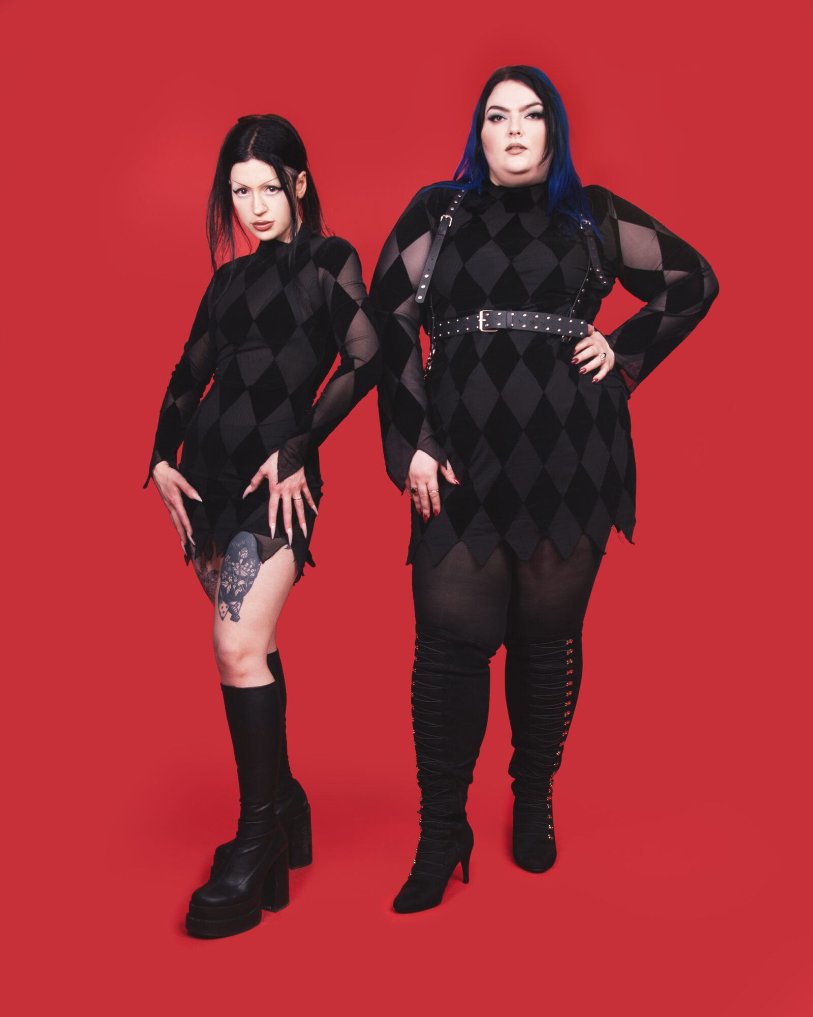 Two women in black argyle-patterned outfits pose against a red background, one with long dark hair and the other with blue hair.