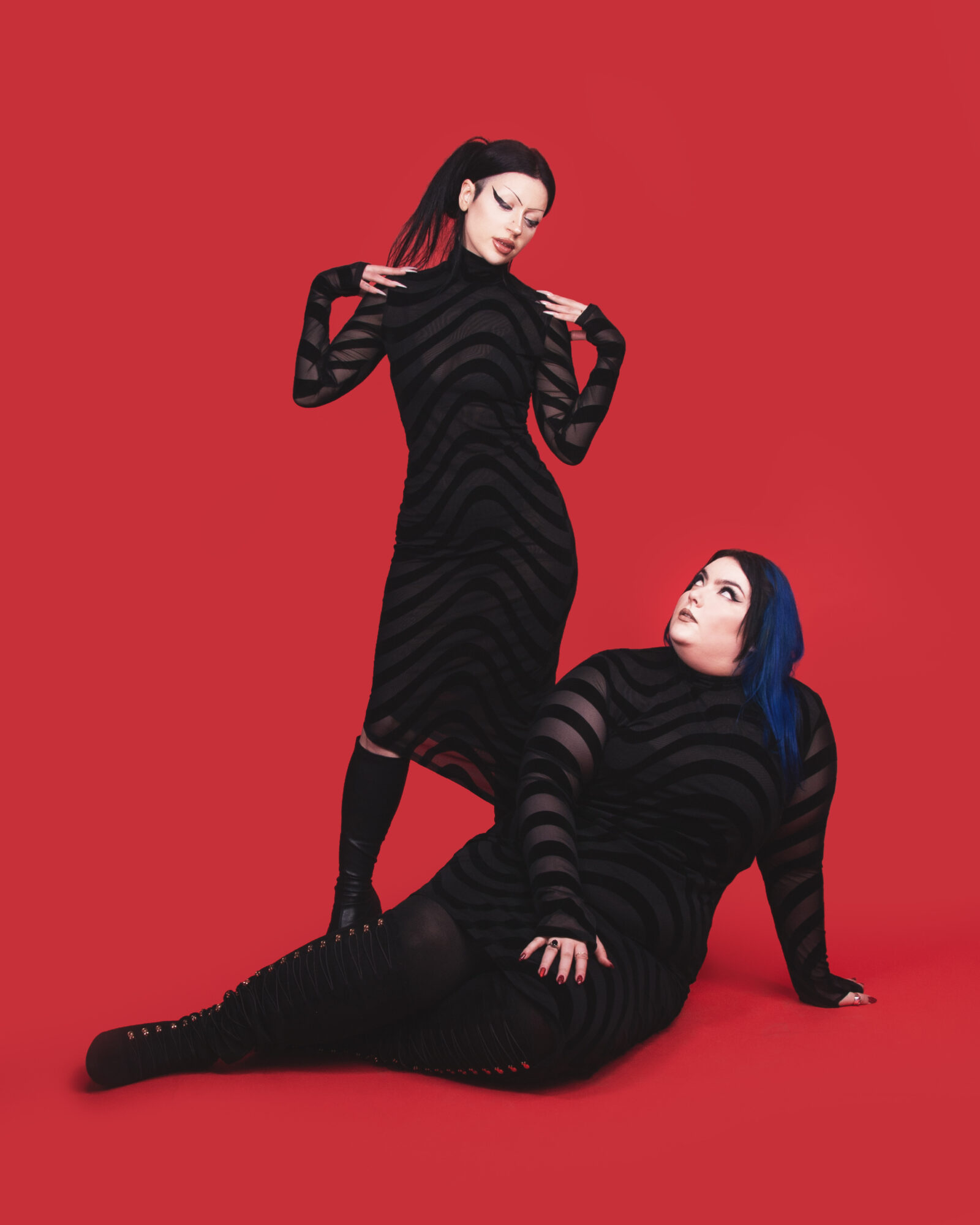 Two women in black outfits with striped patterns pose against a red background, one standing and one sitting on the floor.