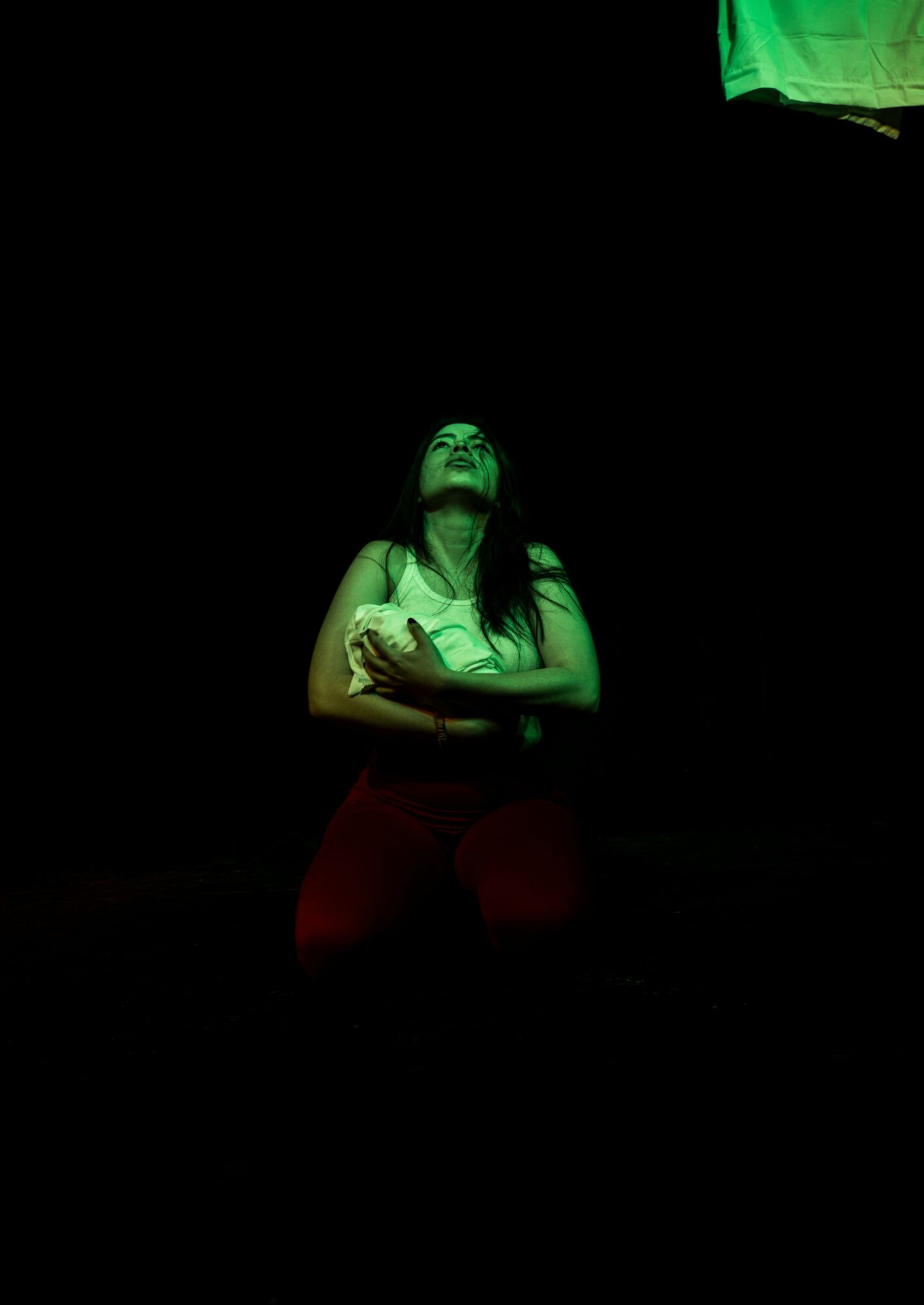 Person with long dark hair looking upward, arms crossed, illuminated by green light against a dark background.