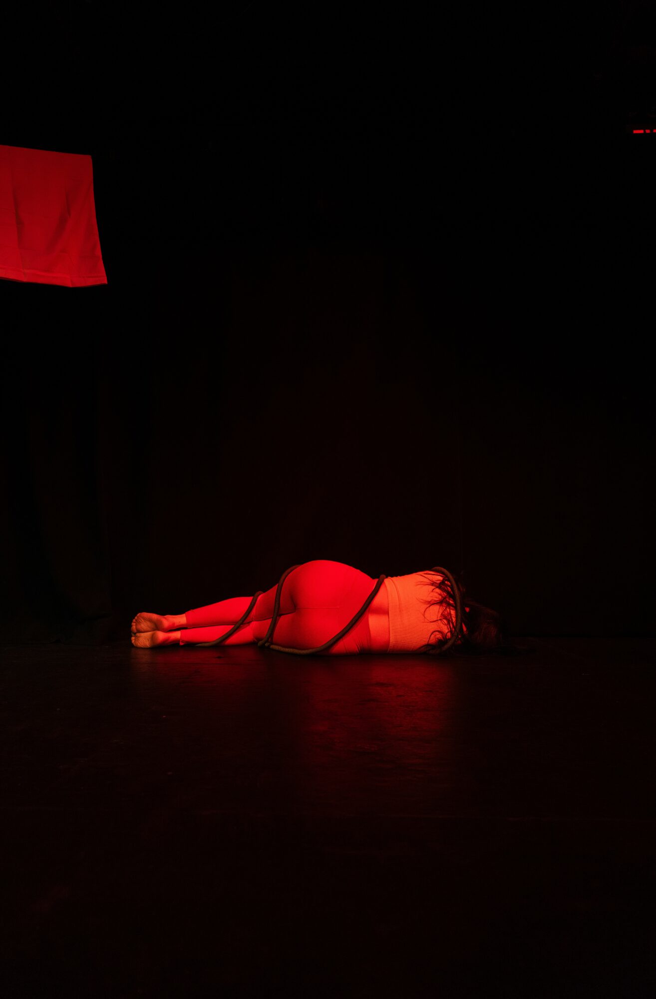 Person lying on stage floor in a dimly lit setting with red lighting, facing downward, with a red cloth nearby.