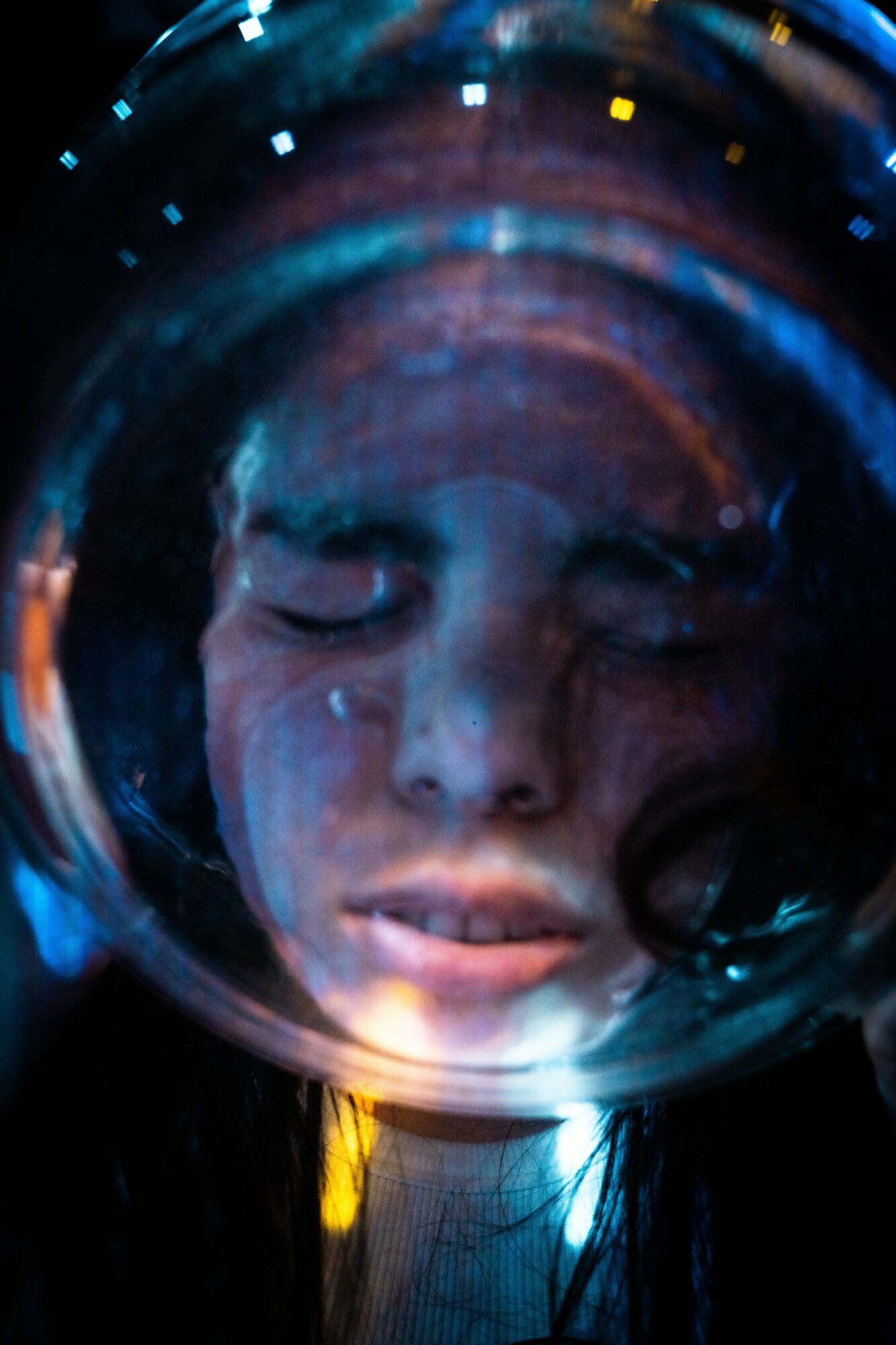 Close-up of a person's face reflected in a circular, glass-like surface with colorful lights, eyes closed, expression neutral.