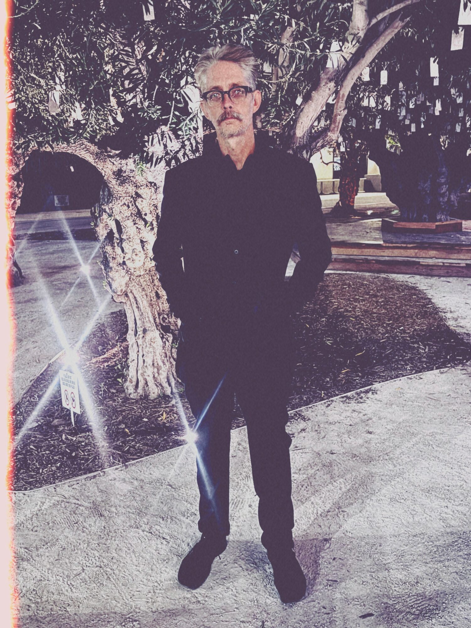 Man with glasses and beard standing outdoors near a tree, wearing dark clothing, with sunlight reflections and a park background.