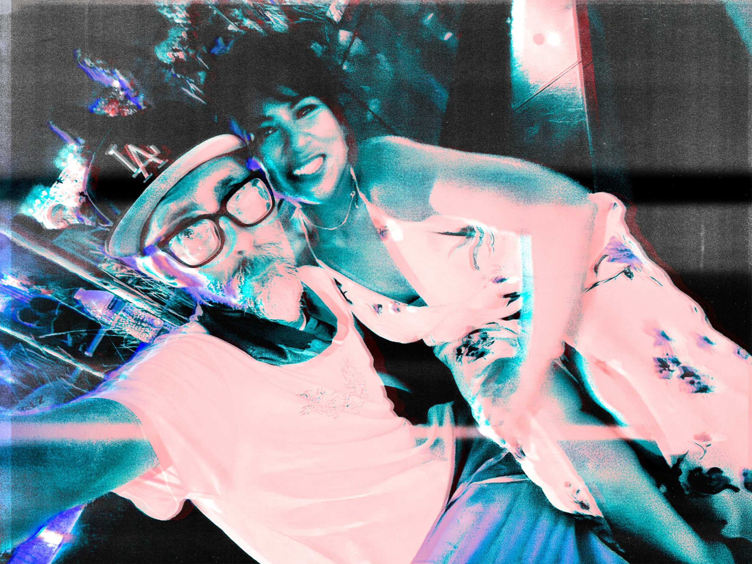 Two people smiling and taking a selfie, one wearing glasses and a hat, the other with short hair, in a colorful setting.