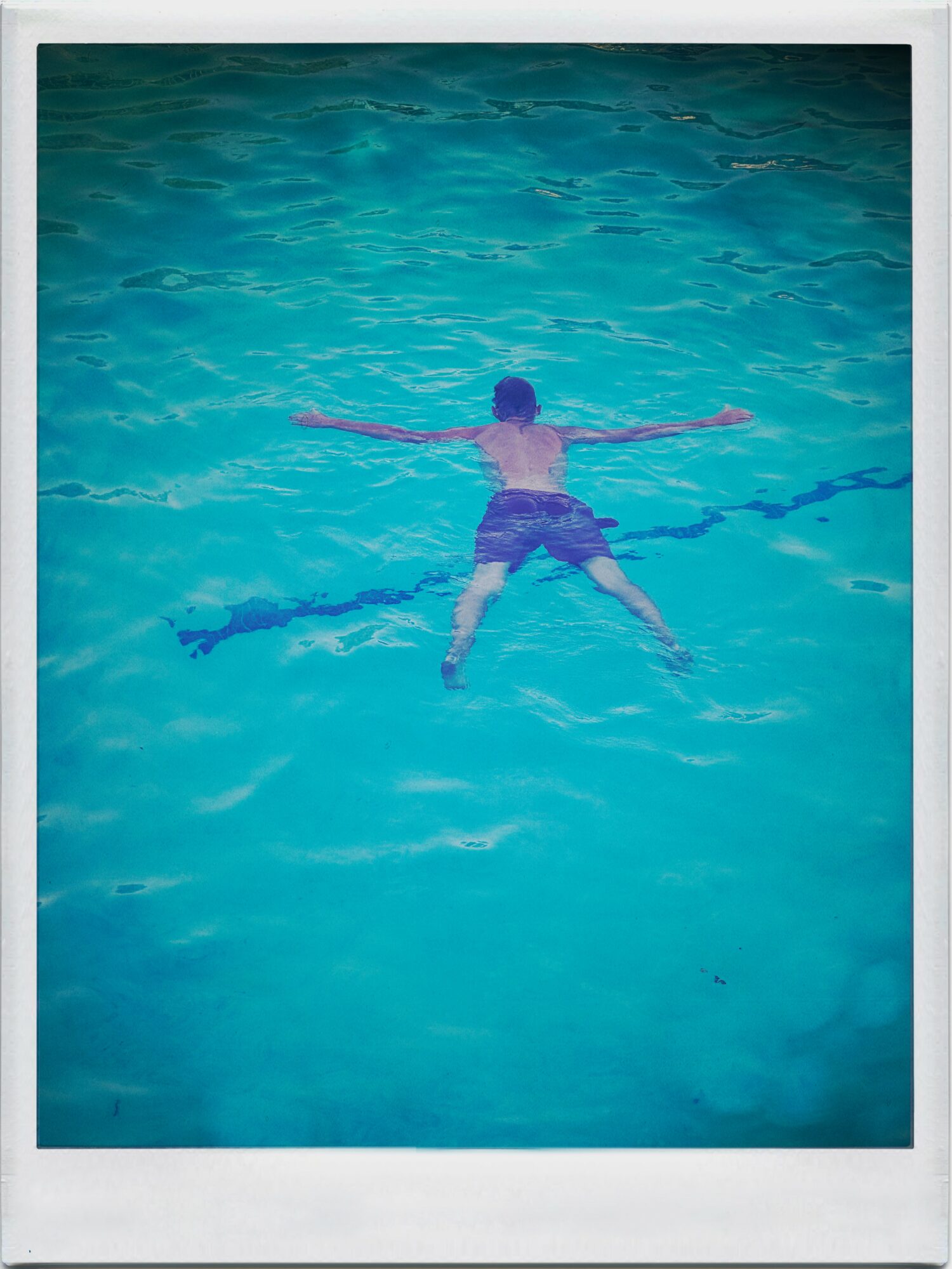 Child floating on back in a swimming pool with arms and legs spread out, wearing a swim cap and shorts.
