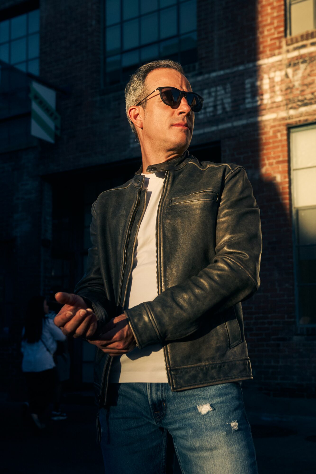 Man wearing sunglasses, leather jacket, and jeans, standing outdoors with buildings in the background.