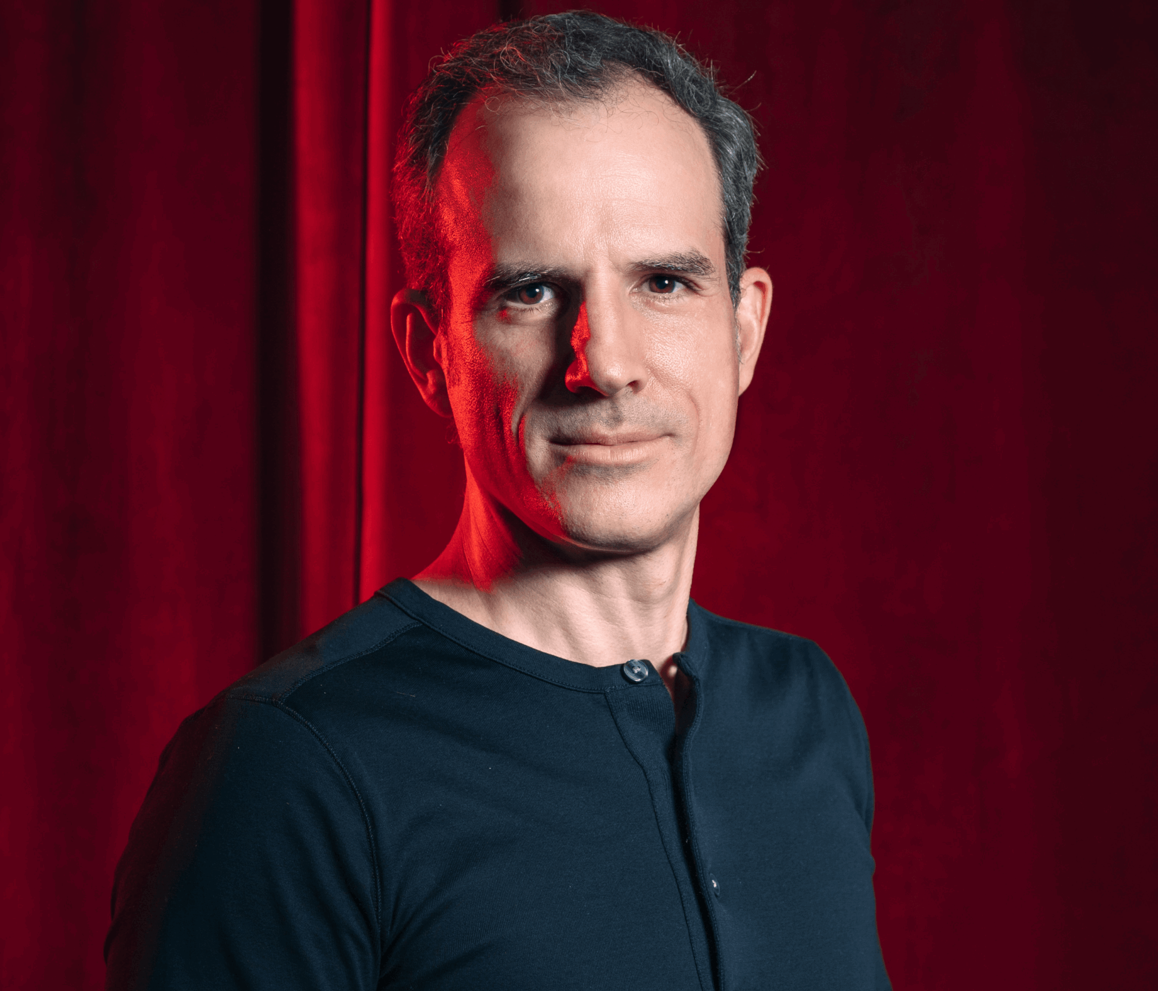 Man with short dark hair and a slight smile, wearing a dark shirt, standing in front of a red curtain background.