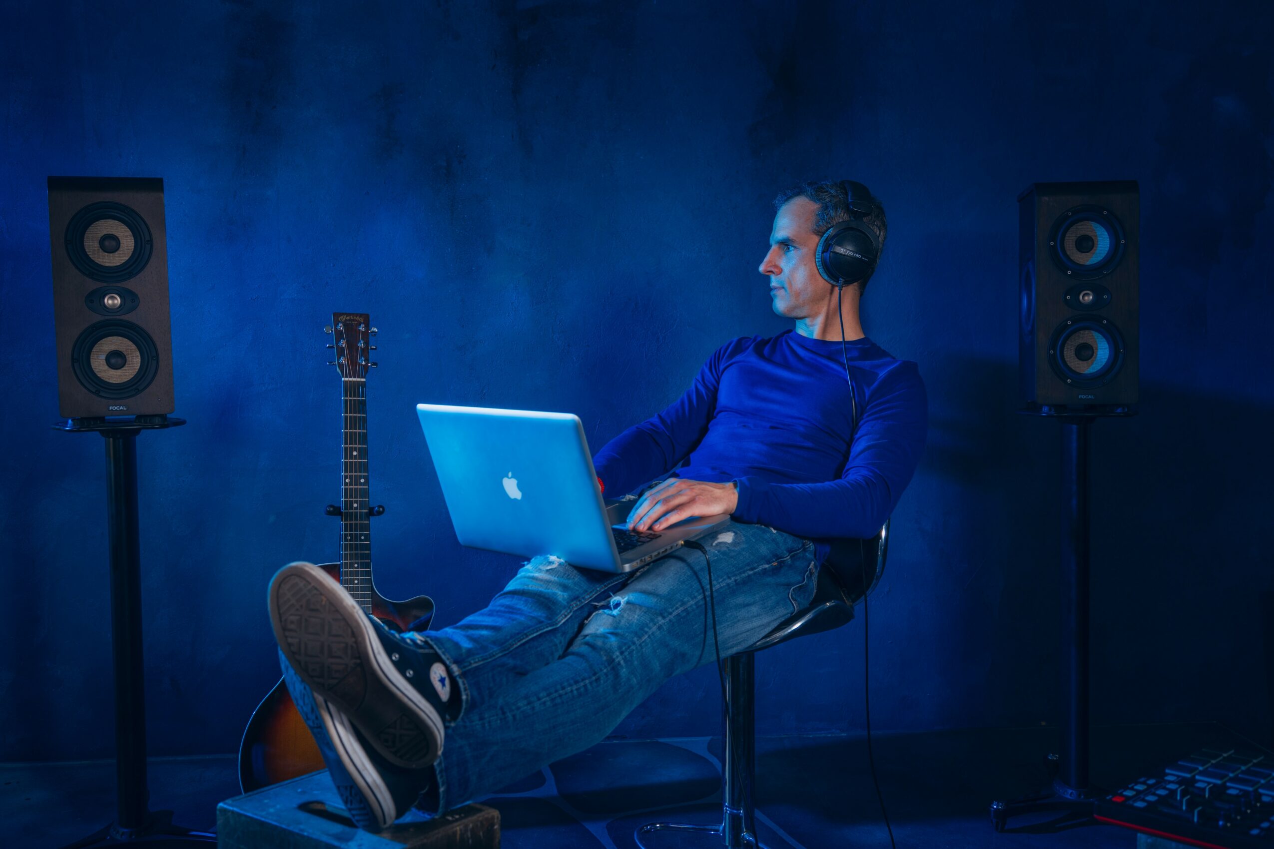 Person sitting with headphones, using a laptop, in a music studio with speakers and guitar, blue background.