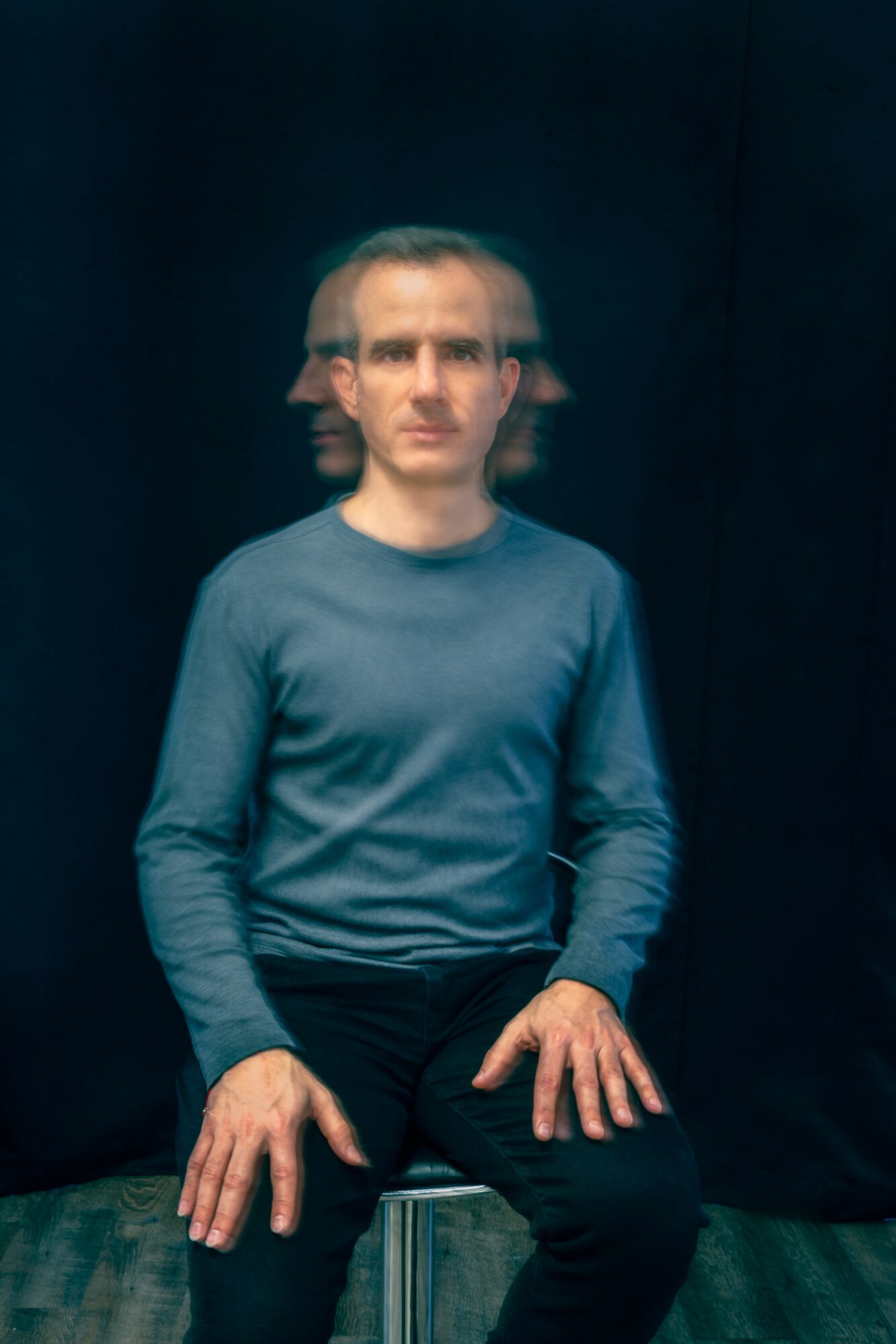 Person with short hair and glasses, sitting with hands on knees, wearing a blue long-sleeve shirt, against a dark background.