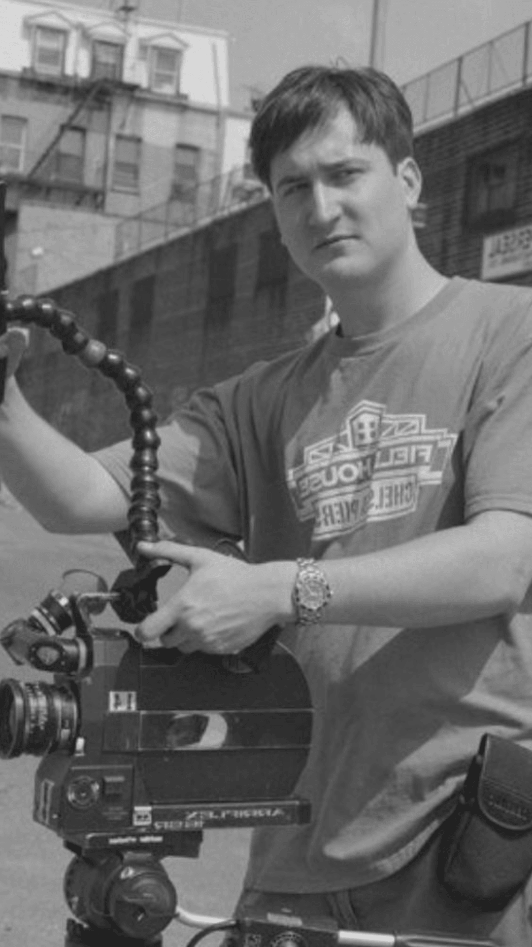 Young man holding a camera with a flexible arm, standing outdoors in front of buildings, wearing a T-shirt and watch.
