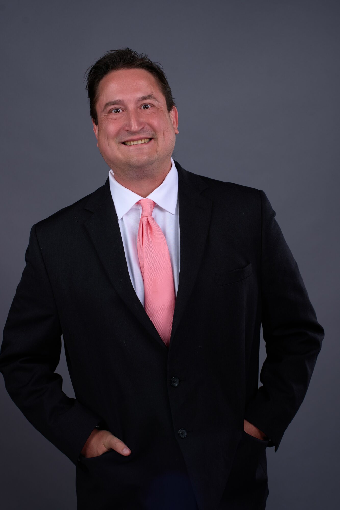 Man in a suit with a pink tie smiling, hands in pockets, dark gray background.