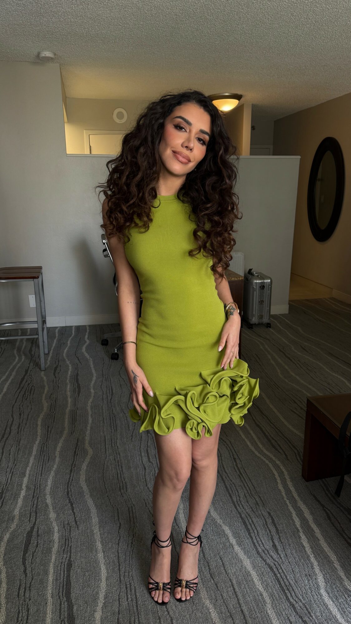 Young woman with curly hair in a lime green dress holding a matching ruffled clutch, standing indoors.