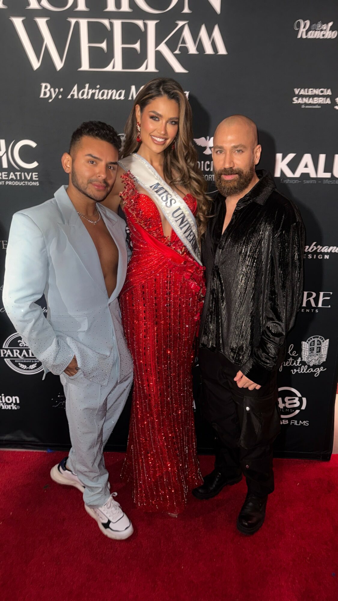 Three people posing on red carpet, woman in red gown with sash, two men in dark and light suits, backdrop with event text