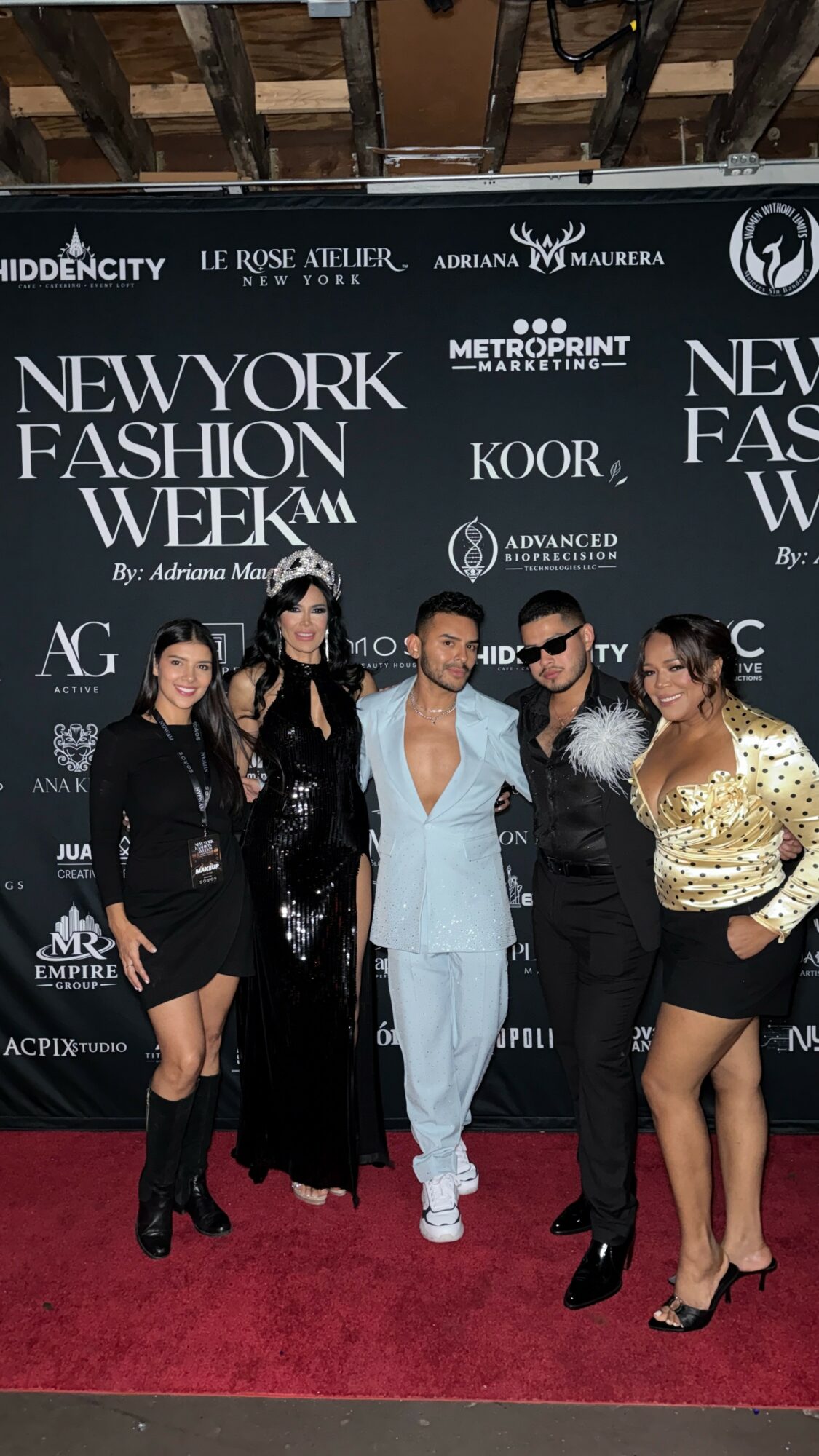 Group of five people posing on red carpet at New York Fashion Week event, with black backdrop and event logos.