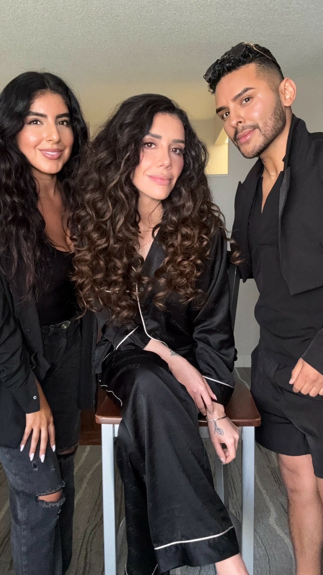 Three people posing indoors, two women and one man, smiling and dressed in dark clothing, with a neutral background.