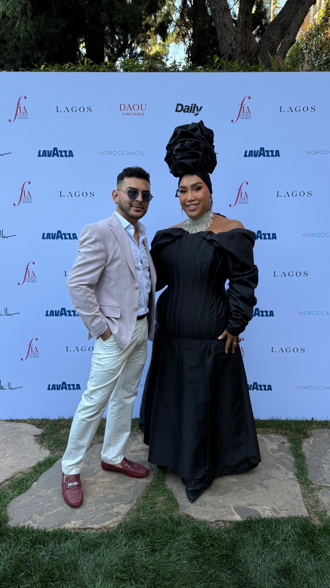 Two people standing outdoors in front of a backdrop with logos, one in a light suit and the other in a black dress with a large headwrap.