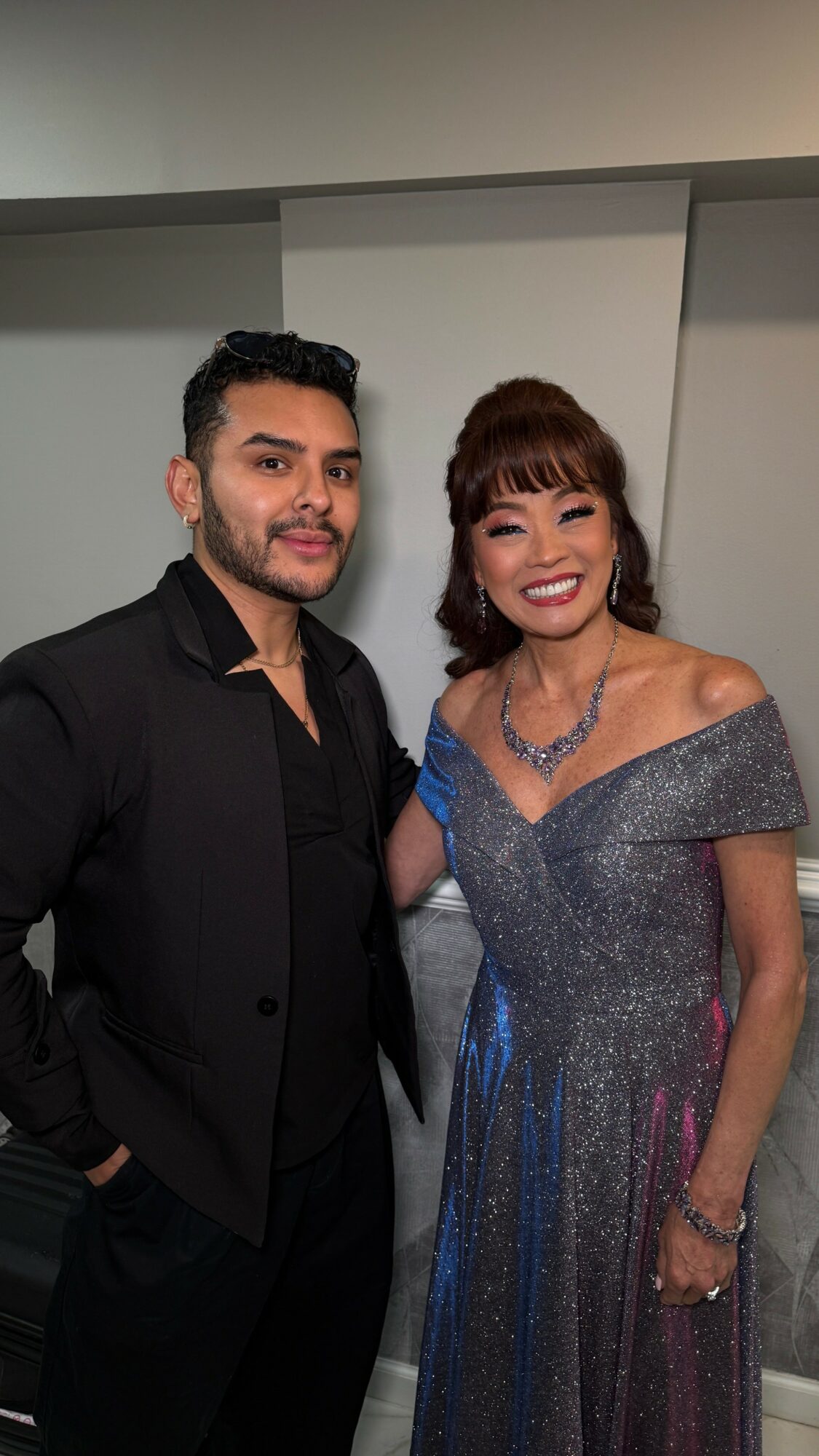 A man and woman dressed formally, smiling, standing indoors with a plain background.