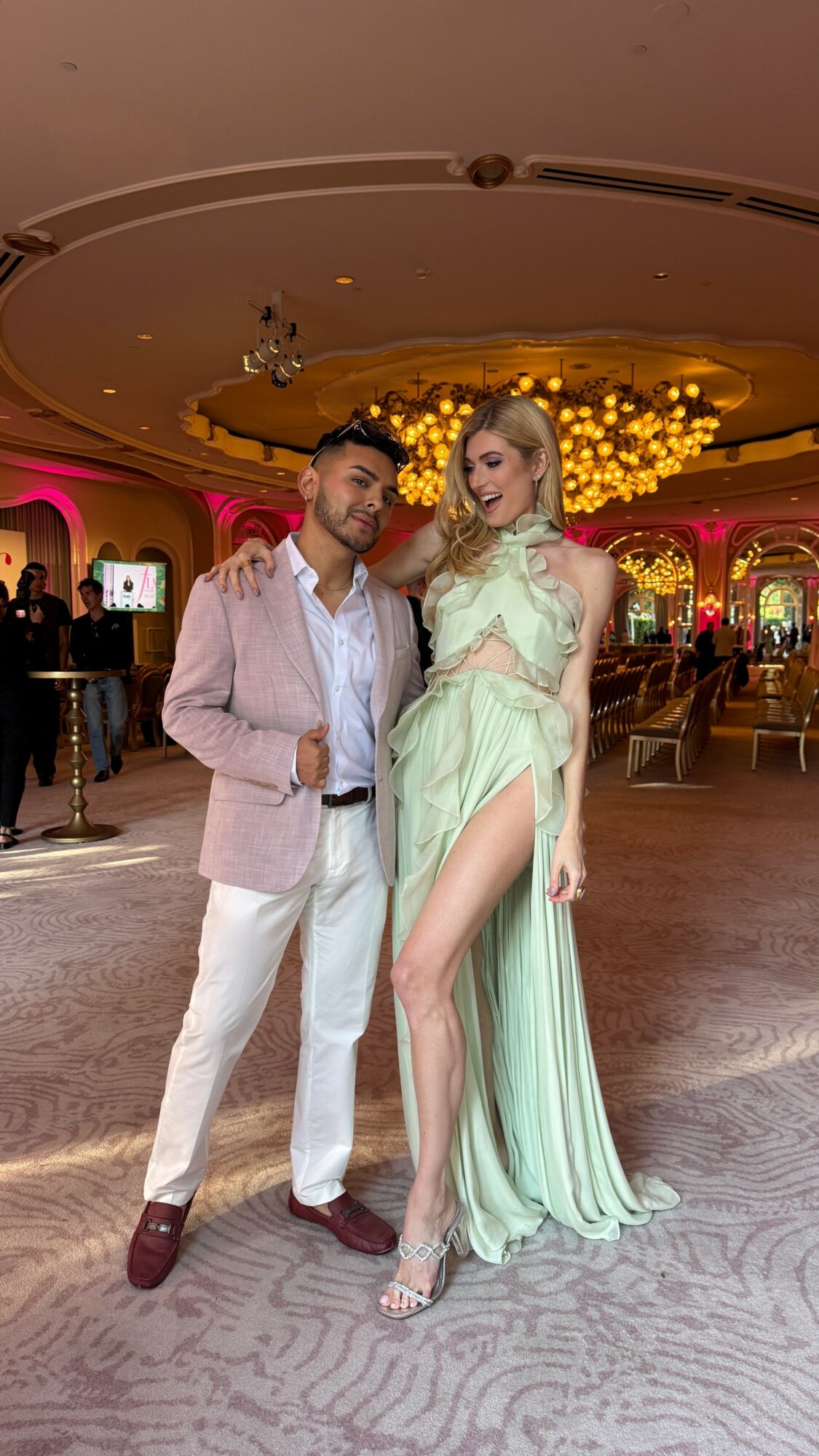 Man and woman pose together in a decorated event space with warm lighting and chandeliers.
