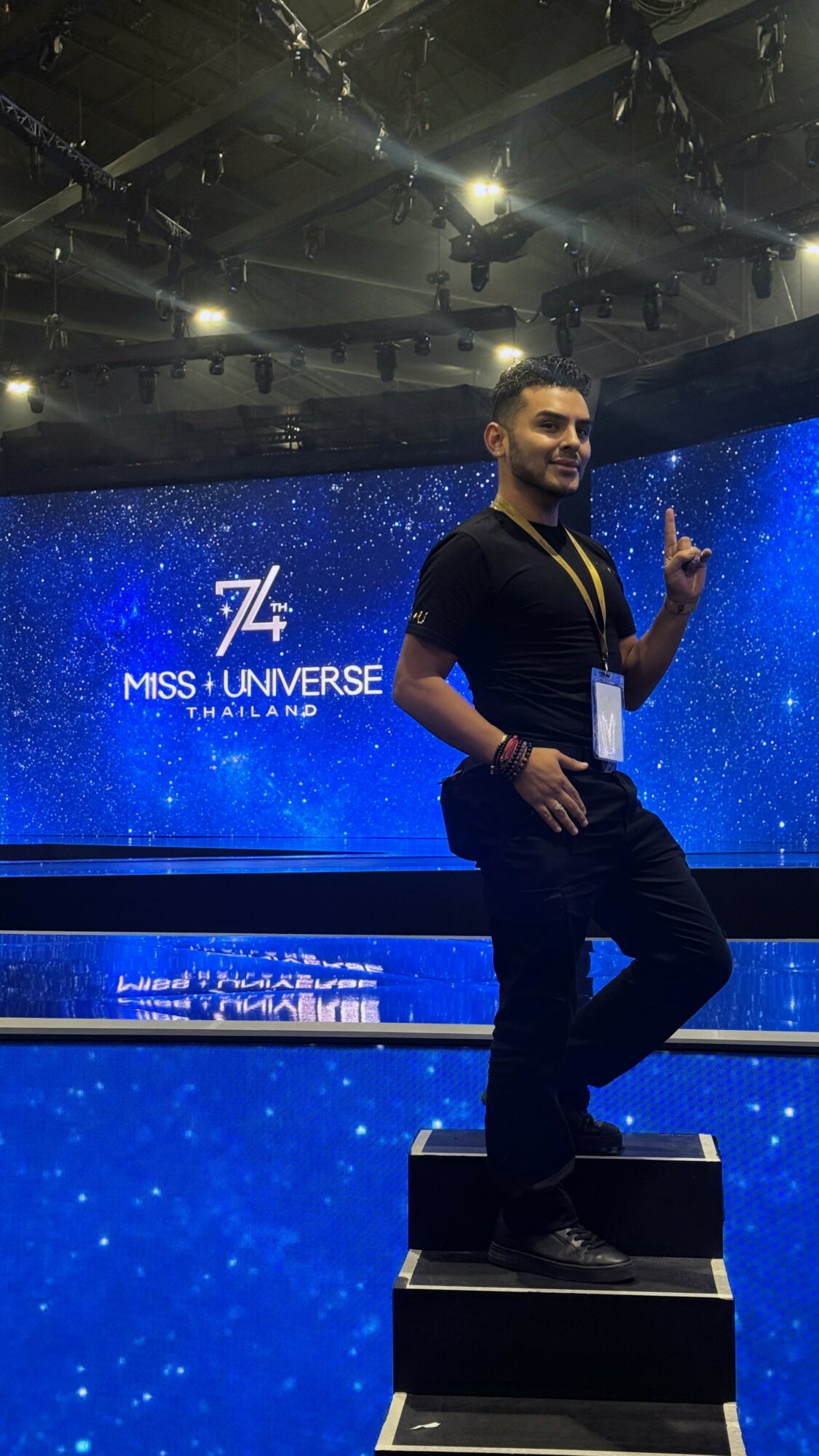 Man standing on a platform in front of a blue starry background with 'Miss Universe Finland' text, smiling and pointing upward.