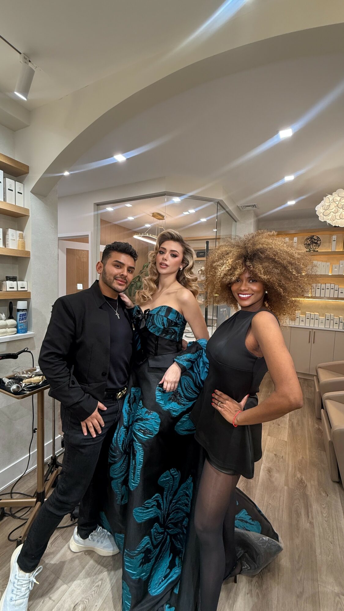 Three people posing indoors, two women and one man, dressed stylishly, smiling, with shelves and lighting in the background.