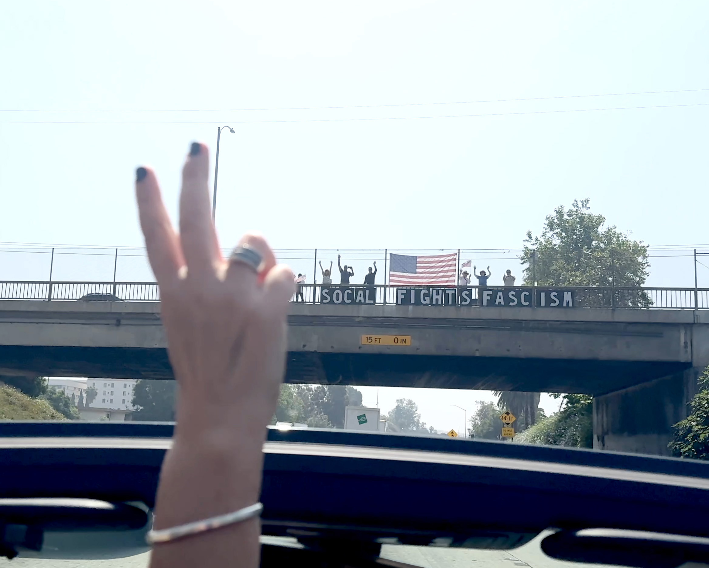 View from inside a vehicle showing a hand with a peace sign, bridge with a banner, and American flag in the background.