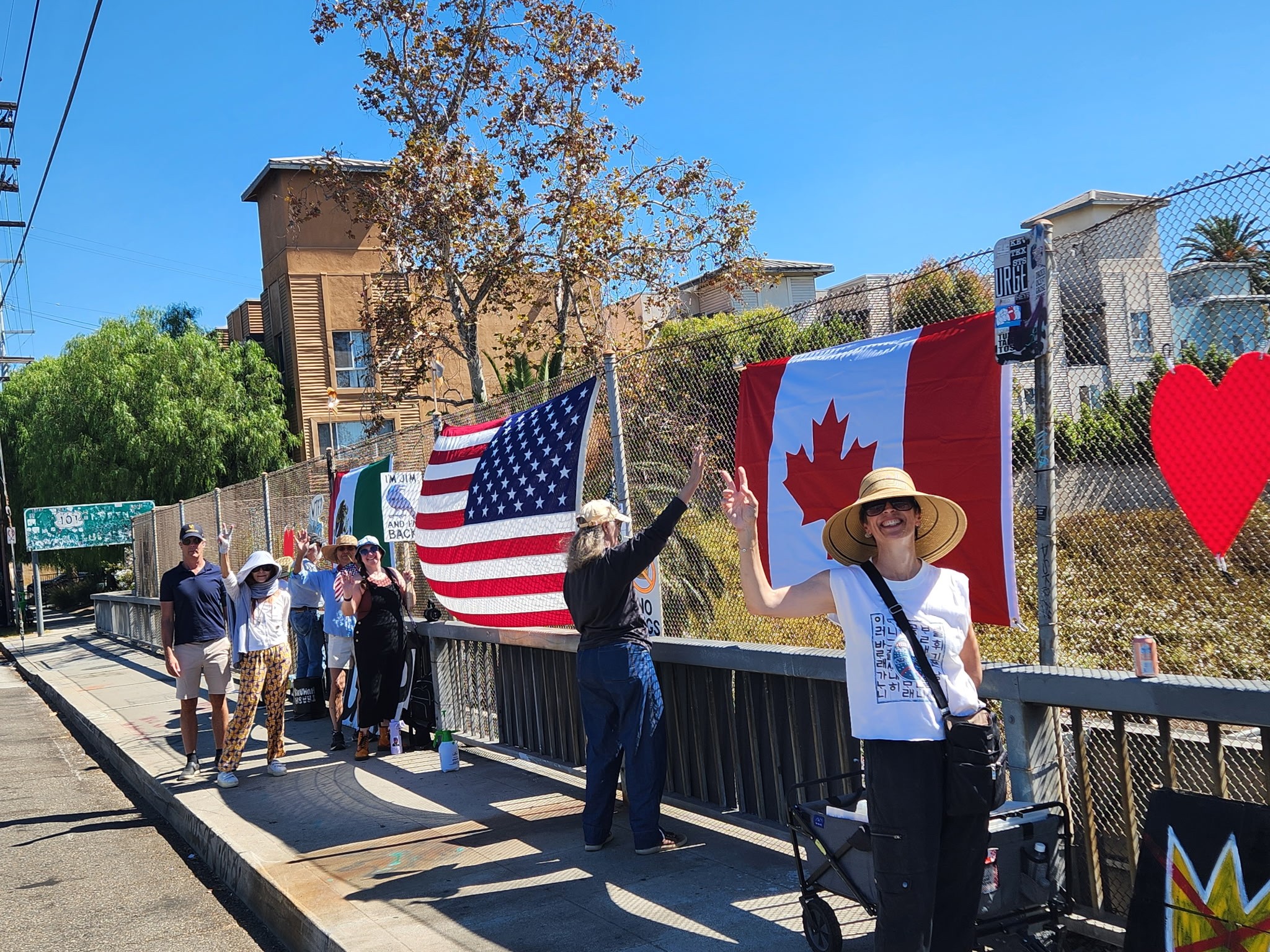 People stand along a fence holding flags, including the United States and Canadian flags, with a building and trees in the background.