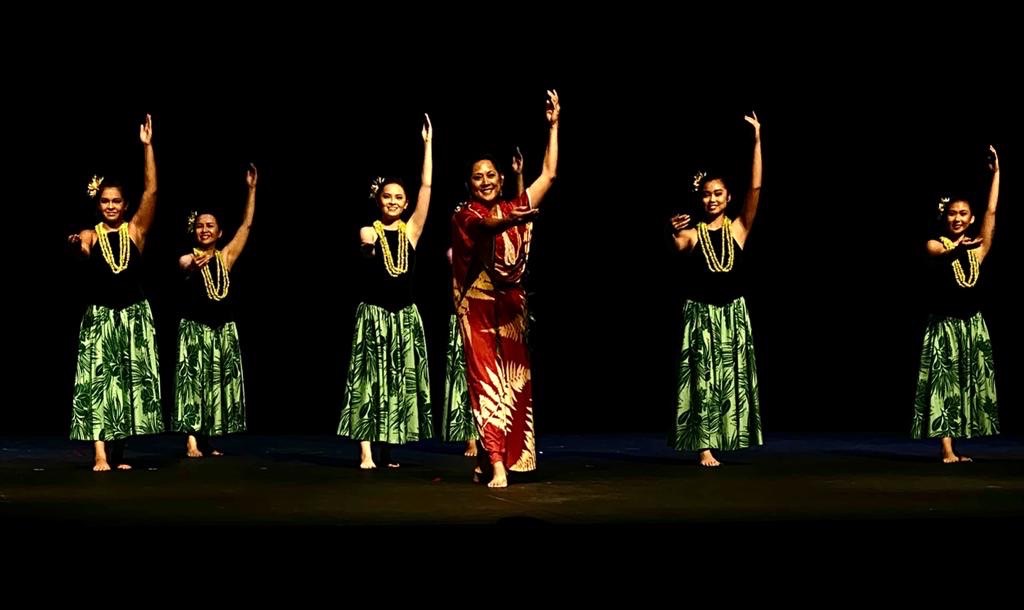 Six dancers on stage, five women in green skirts and one woman in a red dress, all raising one arm, dark background
