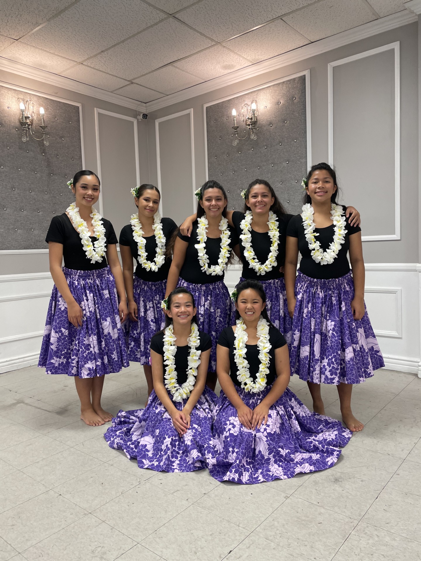Seven women in matching purple skirts and black tops with white floral leis pose in a decorated room, some standing and some sitting.
