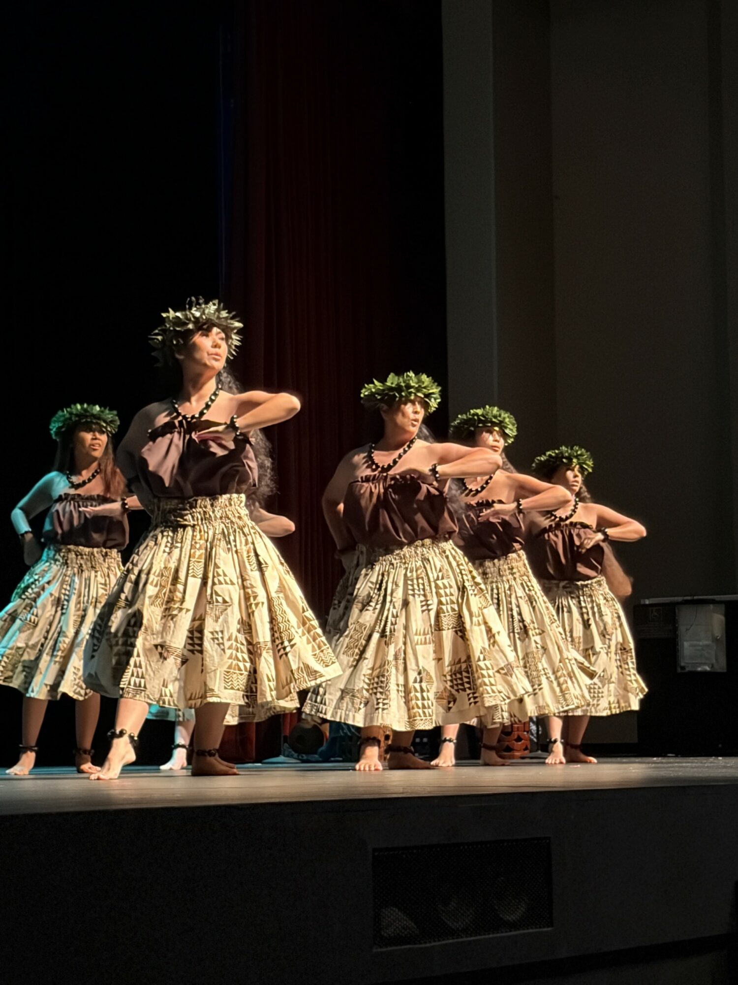 Group of dancers performing on stage, wearing floral crowns and patterned skirts, with arms extended in dance poses.
