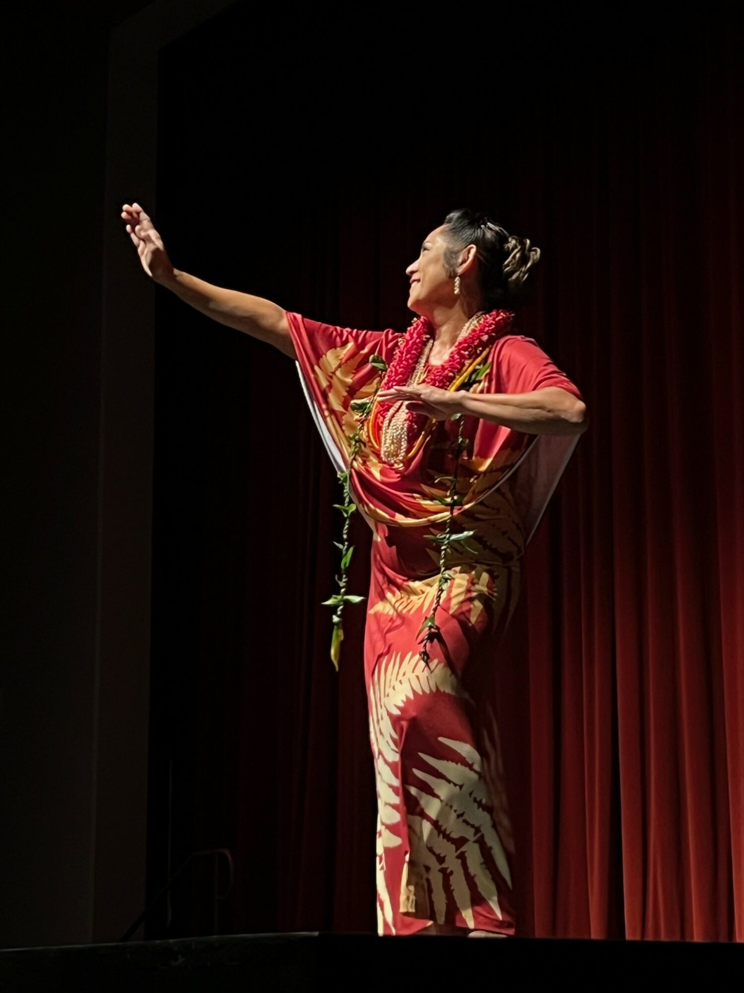 Performer in traditional attire on stage with red curtain, arms extended, expressive pose, hair styled up, colorful costume.