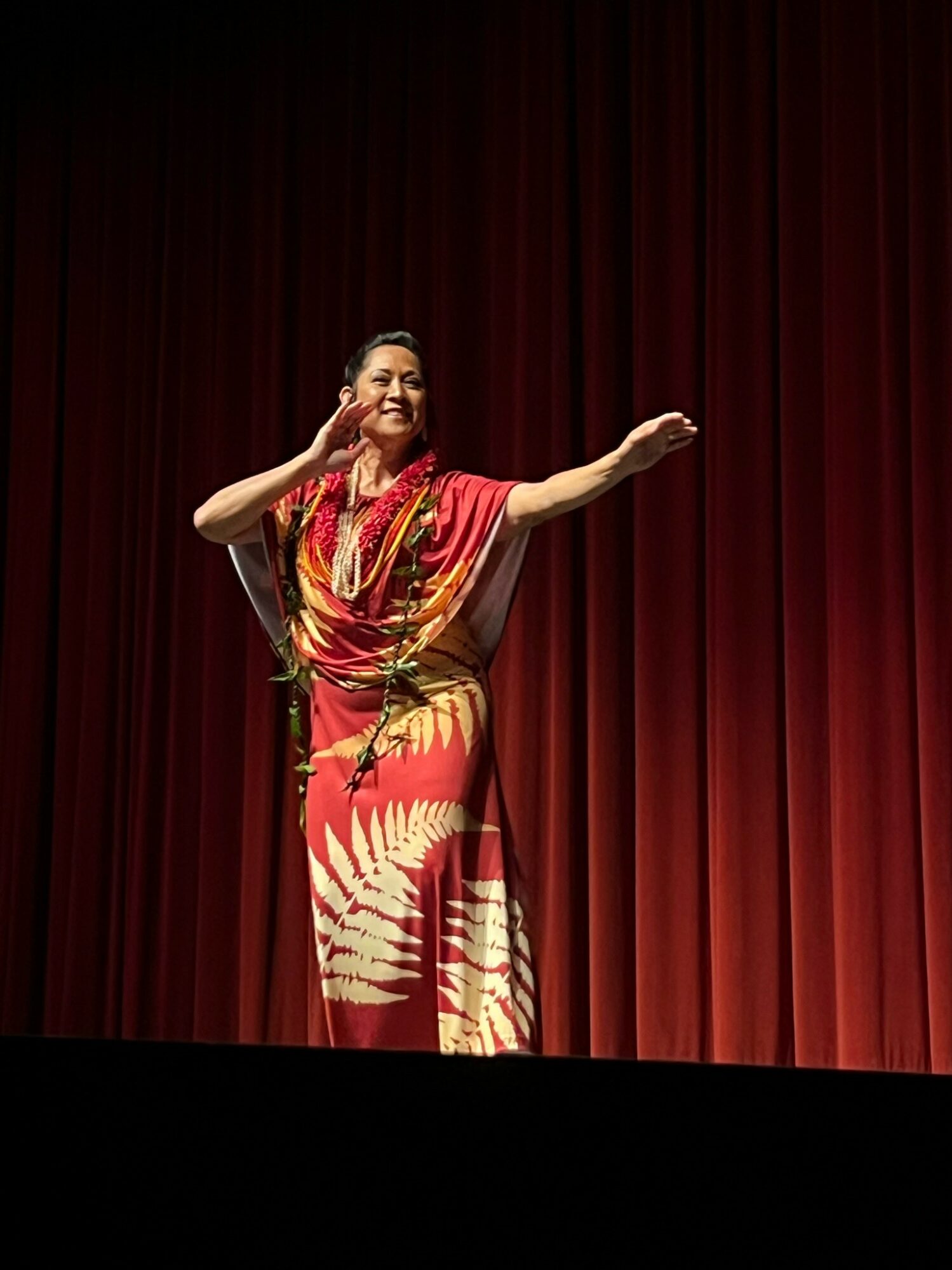 Woman performing on stage with red curtain background, wearing a colorful dress and jewelry, smiling and gesturing with her right hand.
