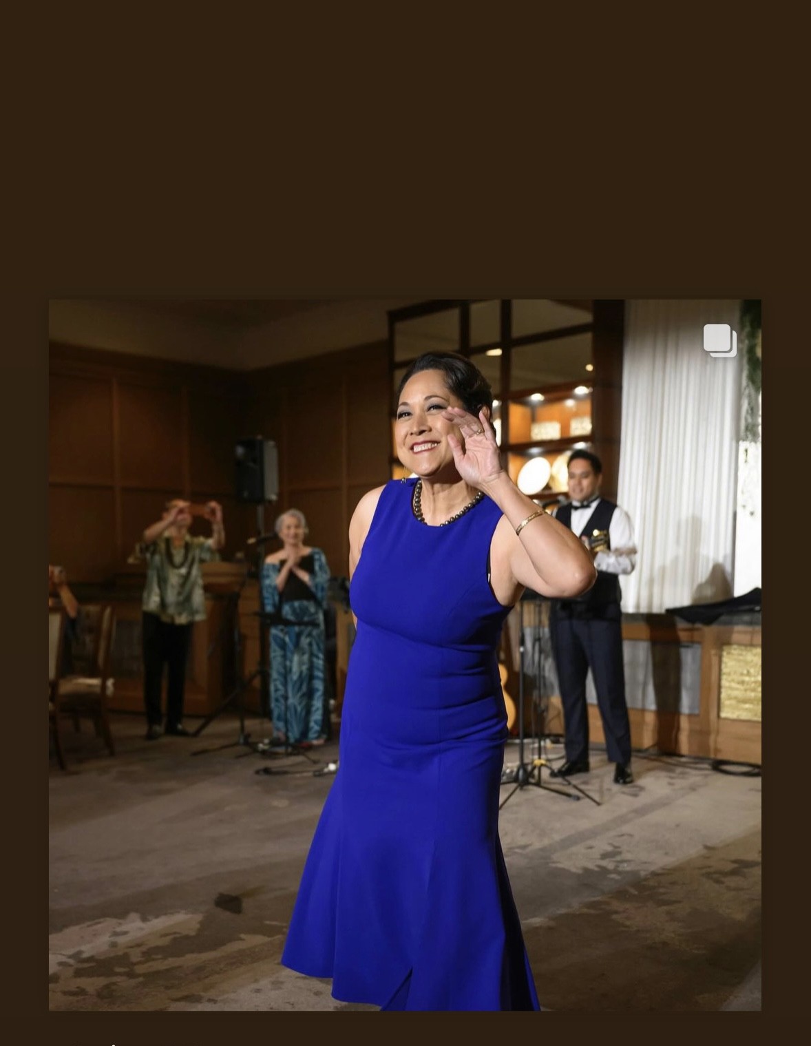 Woman in a blue dress smiling and waving at a celebration with people in the background.