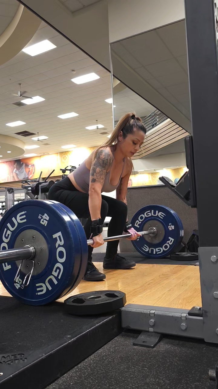 Woman lifting a barbell with weights in a gym, focused on her workout, tattoos visible on her arm.