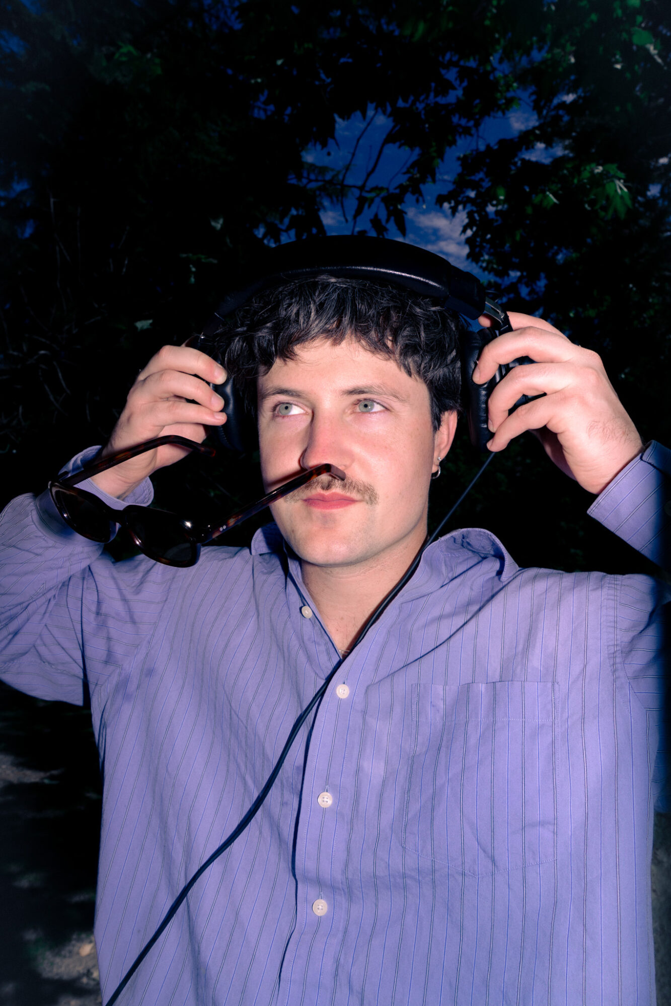Man with dark hair and mustache wearing a blue shirt, holding headphones, outdoors at night with trees in background.