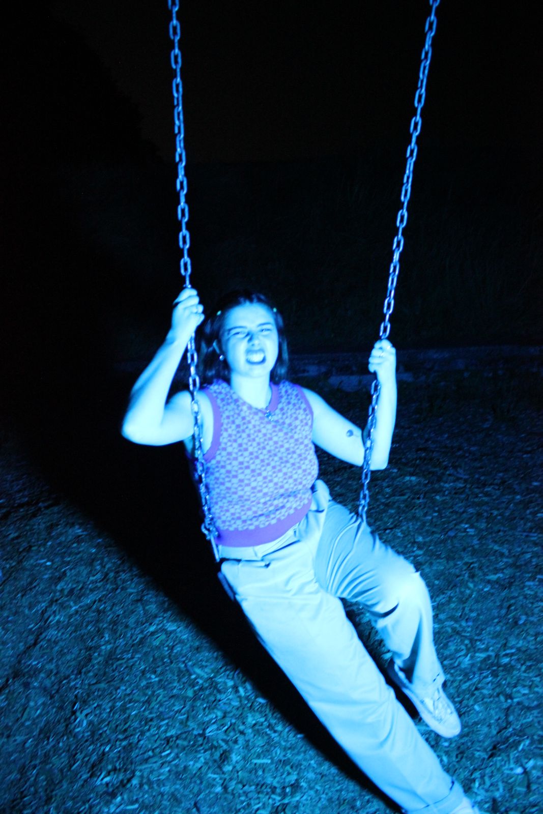 Person sitting on a swing at night, smiling, illuminated by blue light, holding the chains, grass visible below.